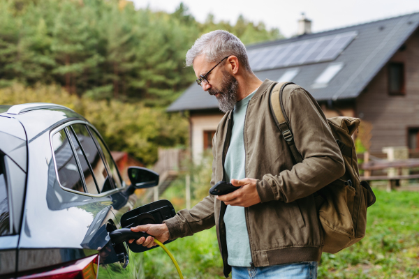 Man charging electric car in front of his house, plugging the charger into the charging port and holding smartphone. House with solar panel system on roof behind him.