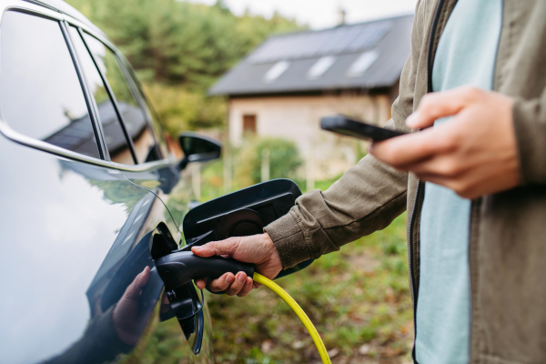 Man charging electric car in front of his house, plugging the charger into the charging port and holding smartphone. House with solar panel system on roof behind him.