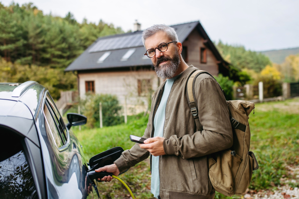 Portrait of a man charging his electric car in front of his house, plugging the charger into the charging port. The man is unplugging the charger from the fully charged car before going to the office.