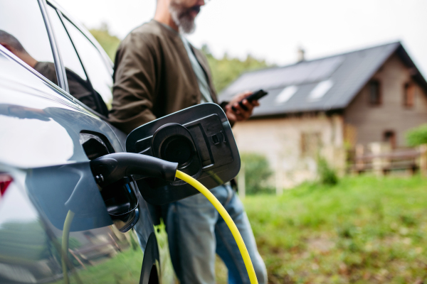 Close up of charging electric car in front of house, charger in charging port. House with a solar panel system on roof behind.