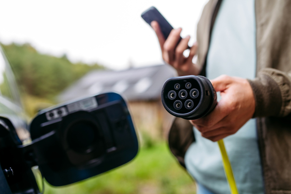 Man charging electric car in front of his house, plugging the charger into the charging port and holding smartphone. House with solar panel system on roof behind him.