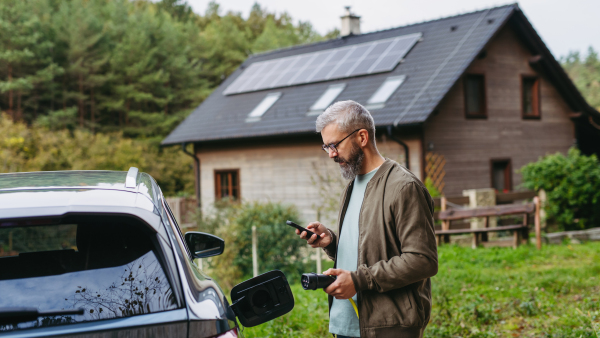 Man charging electric car in front of his house, plugging the charger into the charging port and holding smartphone. House with solar panel system on roof behind him.