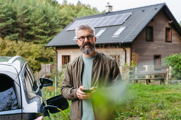 Man charging electric car in front of his house with solar panel system on roof behind him. Man looking at camera and holding smartphone.