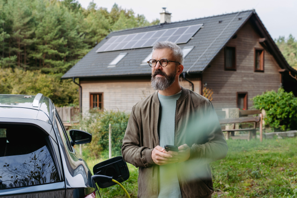 Portrait of a man charging his electric car in front of his house, plugging the charger into the charging port. The man is unplugging the charger from the fully charged car before going to the office.