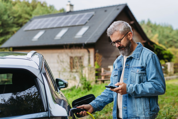 Man charging electric car in front of his house, plugging the charger into the charging port and holding smartphone. House with solar panel system on roof behind him.