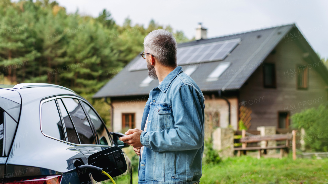 Portrait of a man charging his electric car in front of his house, plugging the charger into the charging port. The man is unplugging the charger from the fully charged car before going to the office.