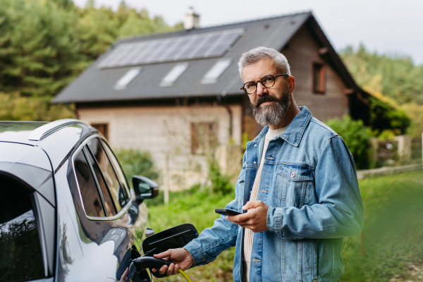 Man charging electric car in front of his house, plugging the charger into the charging port and holding smartphone. House with solar panel system on roof behind him.