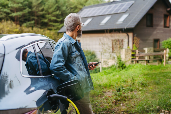 Man charging electric car in front of his house with solar panel system on roof behind him. Mam holding smartphone.