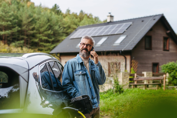 Man charging electric car in front of house while making phone call. House with solar panel system on roof behind him.