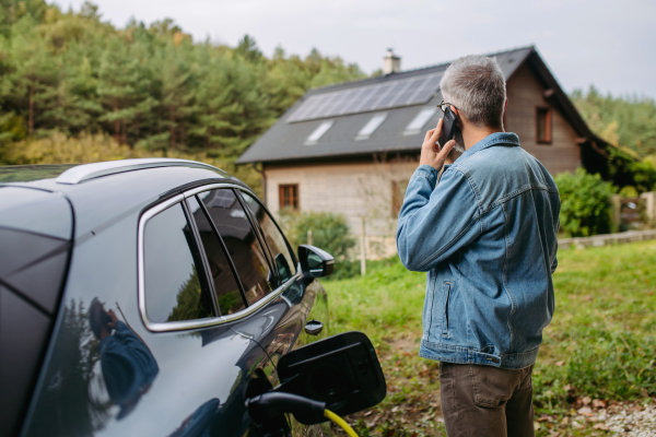 Man charging electric car in front of house while making phone call. House with solar panel system on roof behind him.