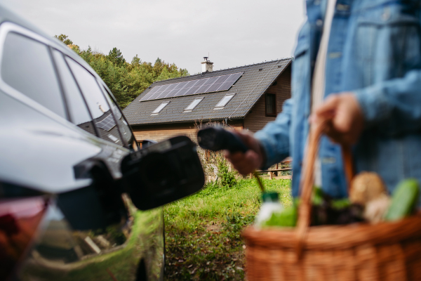 Close up of man holding basket with groceries, plugging charger into eletric car. House with solar panel system on roof behind.