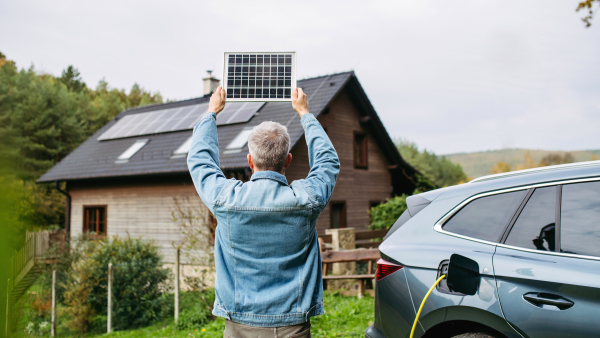 Man holding model of solar panel in hands. Charging electric car and house with solar panel system on roof behind him. Sustainable living.