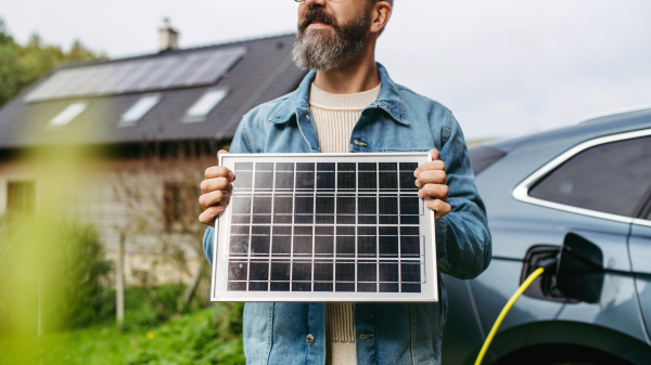 Man holding model of solar panel in hands. Charging electric car and house with solar panel system on roof behind him. Sustainable living.