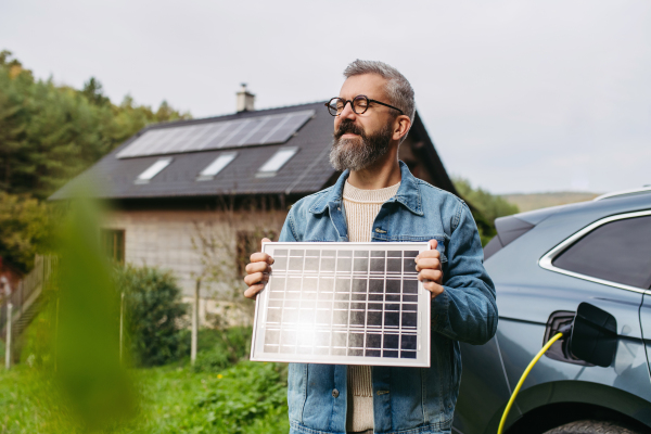 Man holding model of solar panel in hands. Charging electric car and house with solar panel system on roof behind him. Sustainable living.