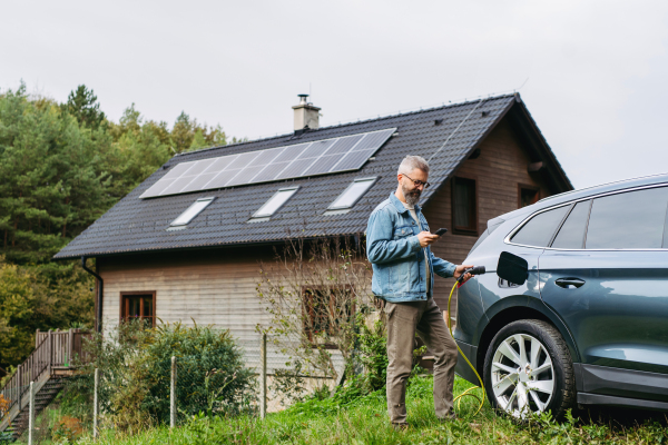 Portrait of a man charging his electric car in front of his house, plugging the charger into the charging port. The man is unplugging the charger from the fully charged car before going to the office.