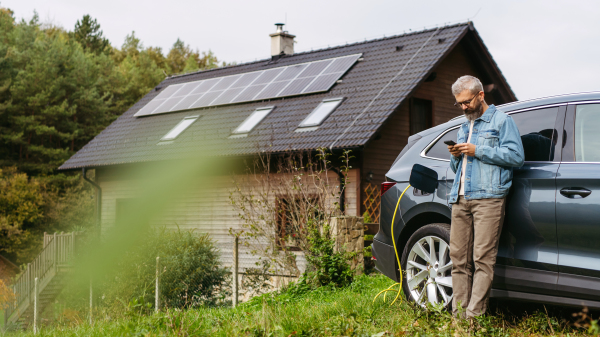 Man charging electric car in front of his house with solar panel system on roof behind him. Man leaning against car while scrolling on smartphone.