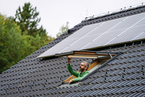 Mature man closing skylight, roof window, solar panels above him. Sustainable lifestyle and green households