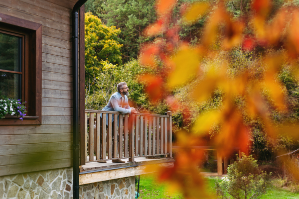 Man is enjoying a relaxing, peaceful moment at home, standing on patio, leaning against railing and looking at the autumn nature around him.