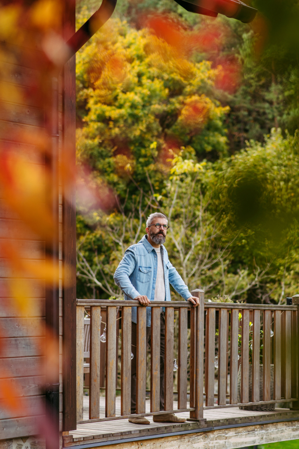 Man is enjoying a relaxing, peaceful moment at home, standing on patio and looking at autumn nature around him.