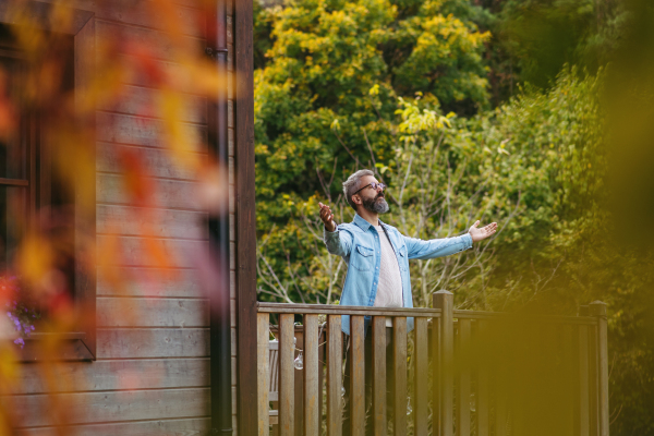 Man standing on patio with outstretched arms and closed eyes, autumn nature around him. Peaceful, relaxing moment at home.