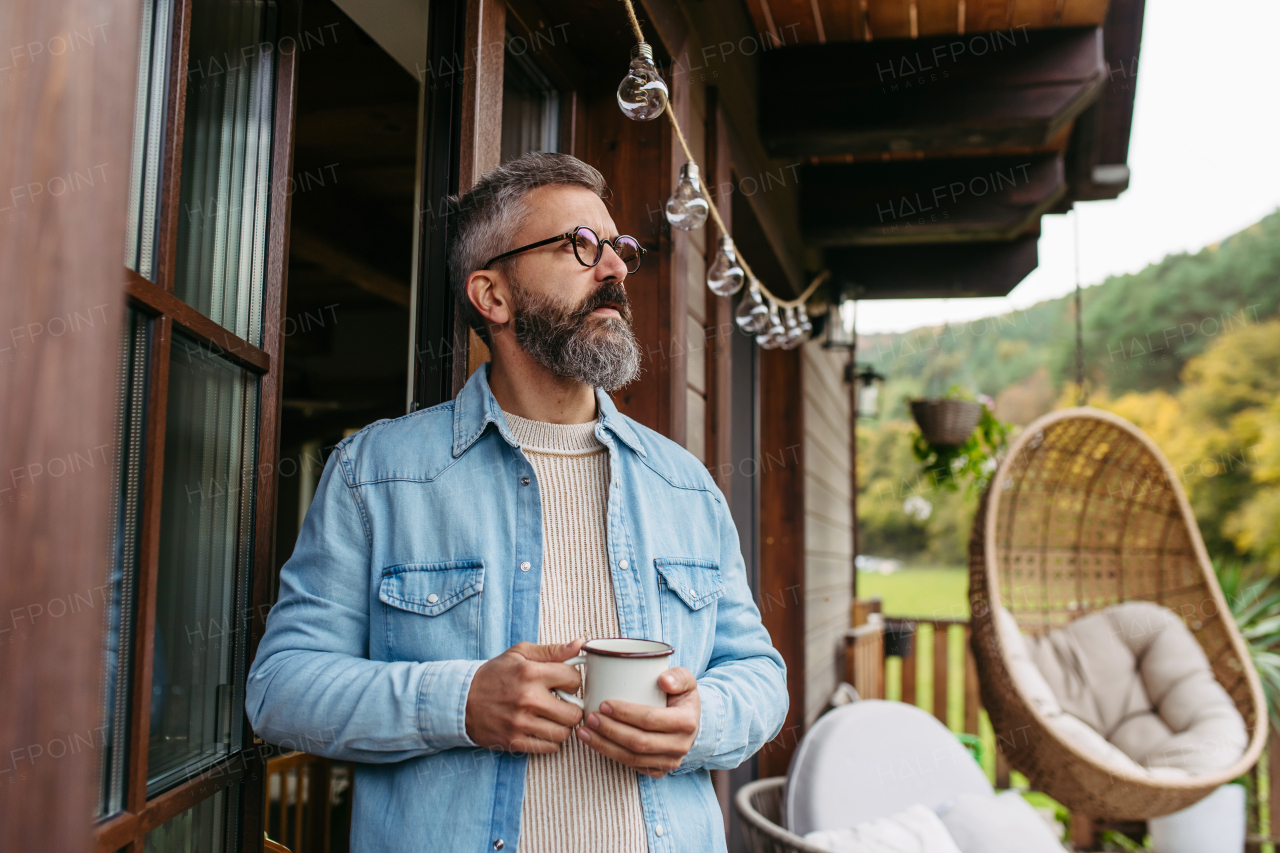 Man is enjoying cup of warm tea, coffee, having relaxing moment at home, standing on patio and looking at autumn nature around