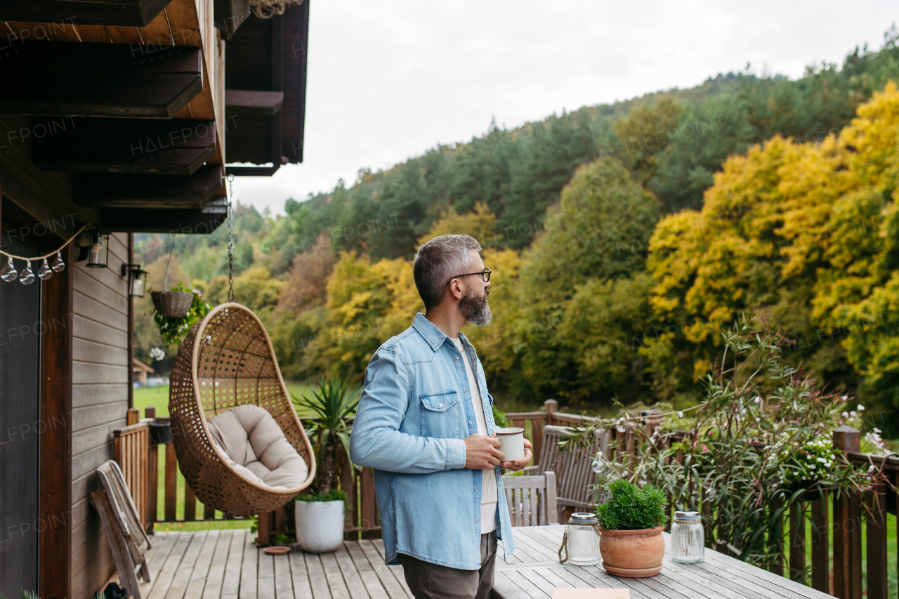 Man is enjoying cup of warm tea, coffee, having relaxing moment at home, standing on patio and looking at autumn nature around