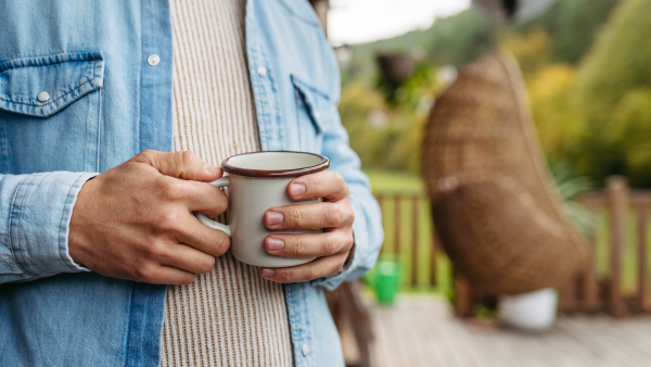 Close up of hands holding cup of warm tea, coffee, having relaxing moment at home, standing on a patio autumn nature around.