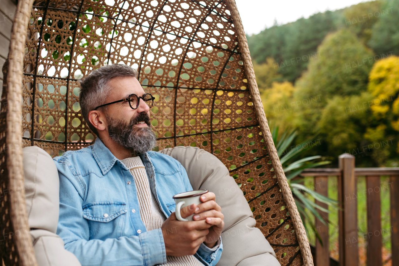 Man is enjoying cup of warm tea, coffee, having relaxing moment at home, sitting in hanging chair on the patio.