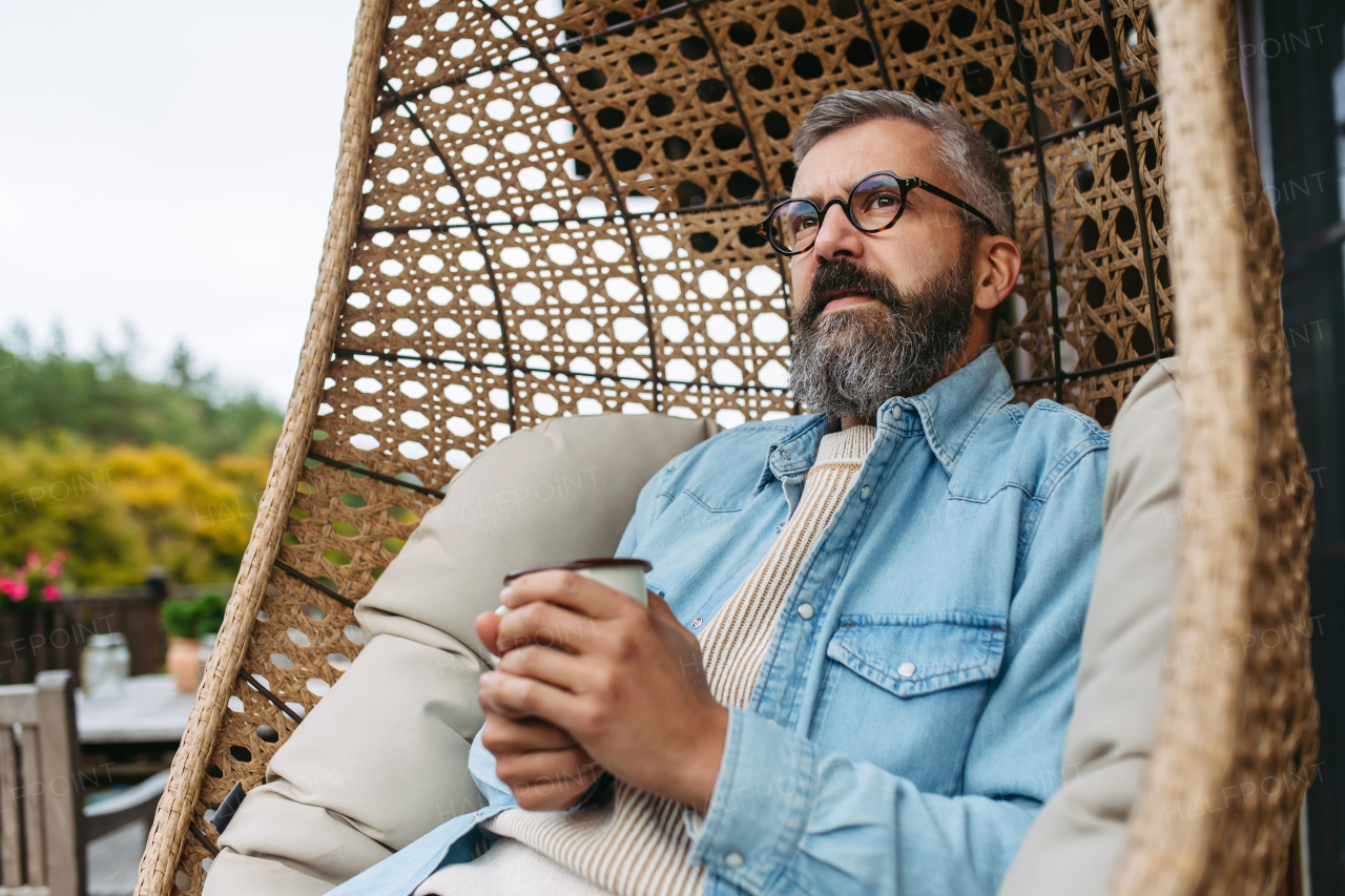 Man is enjoying cup of warm tea, coffee, having relaxing moment at home, sitting in hanging chair on the patio.