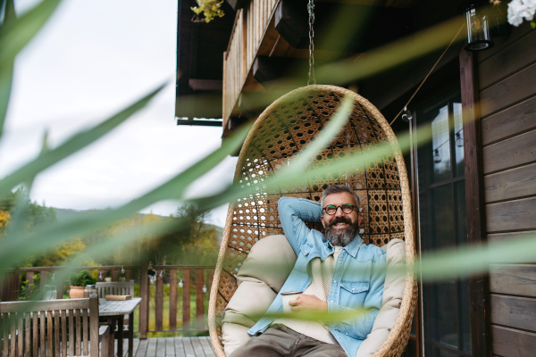 Man is enjoying cup of warm tea, coffee, having relaxing moment at home, sitting in hanging chair on the patio.