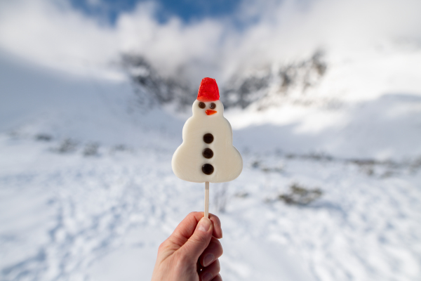 Snowman shaped lollipop held in a hand. In the background beautiful snowy landscape with snow covered mountain peaks.