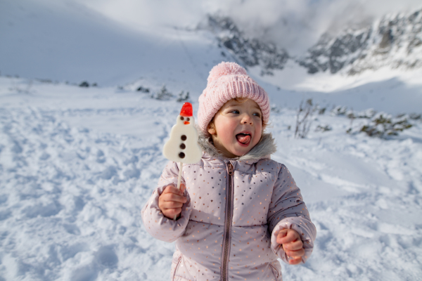 Small girl licking a snowman-shaped lollipop, standing in the middle of snowy nature. Genjoying winter holiday in the mountains with family