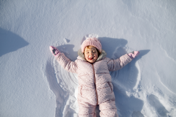 Portrait of a small girl making snow angel, enjoying winter holiday in the mountains with family. Girl lying and playing in snow.