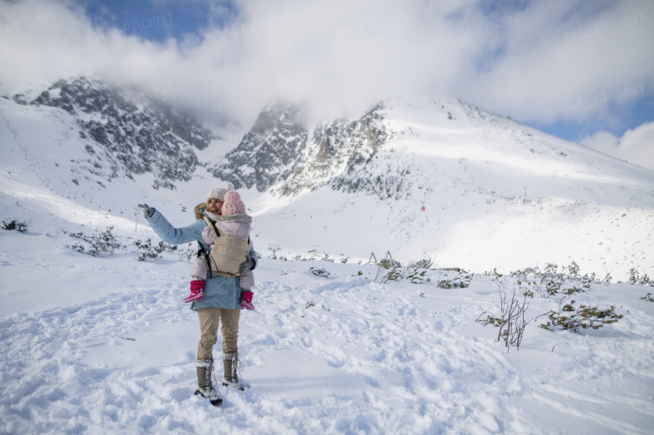 Mother standing in the middle of snowy landscape with daughter in baby carrier. Mom and girl enjoying winter holiday in mountains.