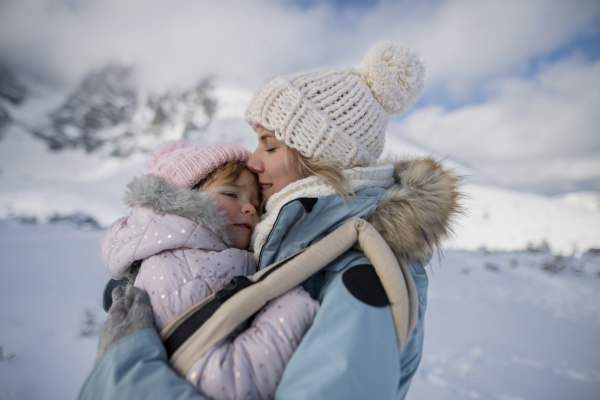 Mother standing in the middle of snowy landscape with daughter in baby carrier. Mom and girl enjoying winter holiday in mountains.