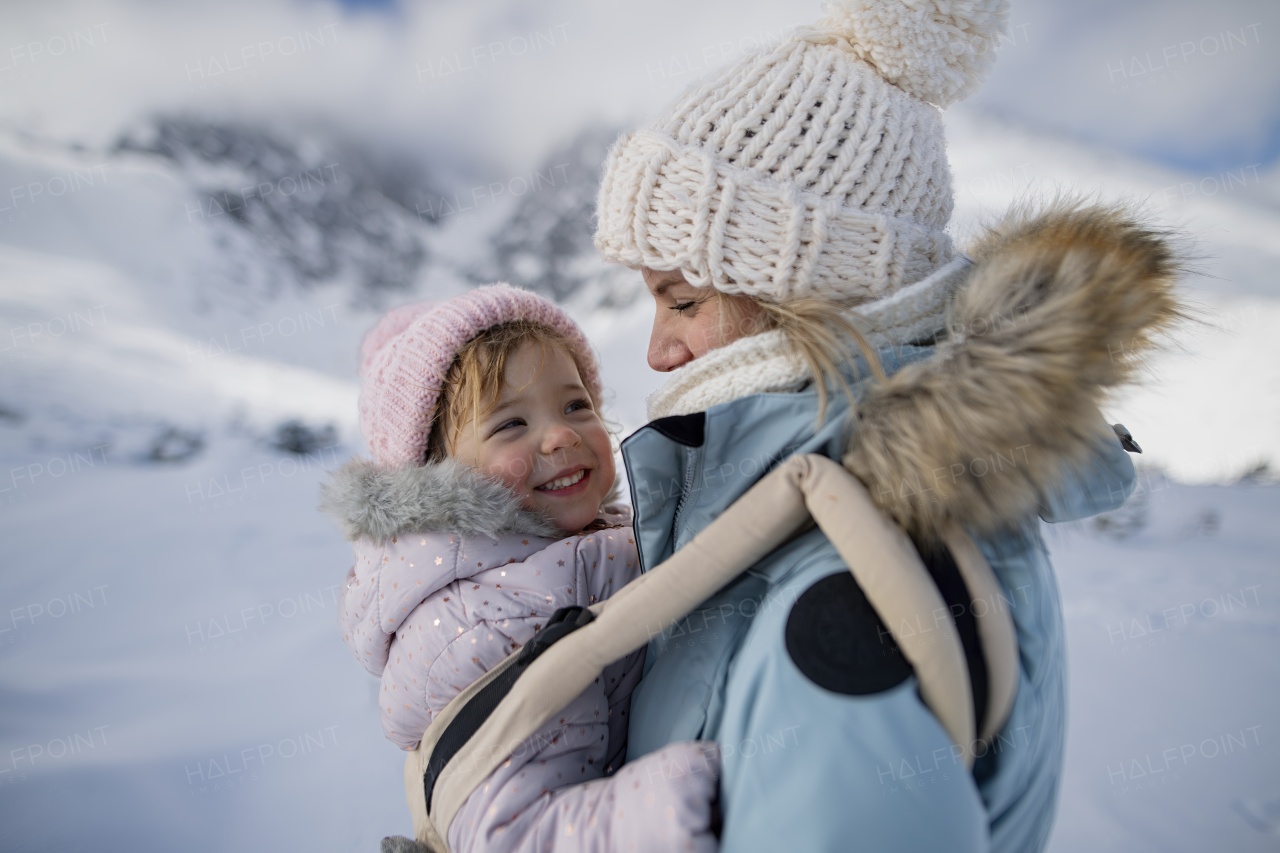 Mother standing in the middle of snowy landscape with daughter in baby carrier. Mom and girl enjoying winter holiday in mountains.