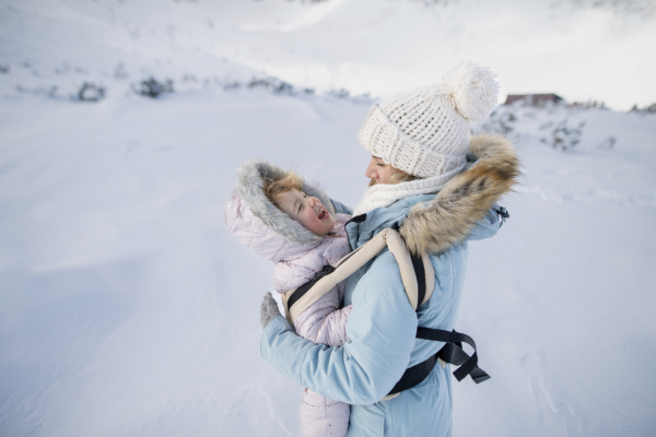 Mother standing in the middle of snowy landscape with daughter in baby carrier. Mom and girl enjoying winter holiday in mountains.