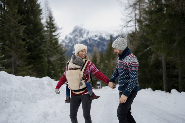 Young family is enjoying winter holiday in the mountains, mother carry small girl in carrier while walking through the snowy forest