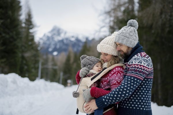 Young family is enjoying winter holiday in the mountains, mother carry small girl in carrier while walking through the snowy forest