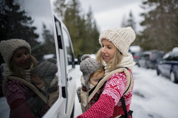 Mother standing in the middle of snowy parking lot with sleeping daughter in baby carrier. Mom and girl enjoying winter holiday in mountains.