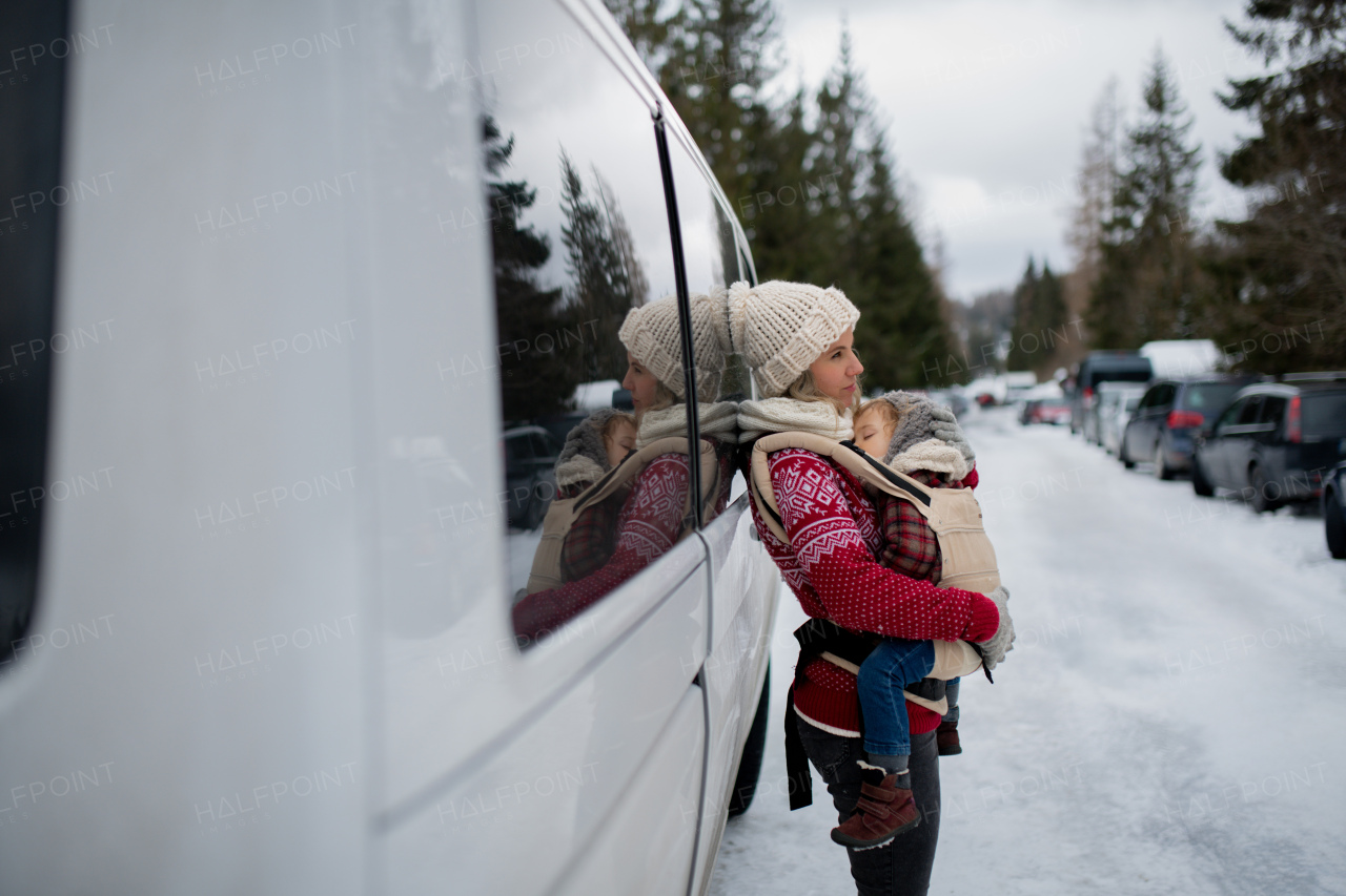 Mother standing in the middle of snowy parking lot with sleeping daughter in baby carrier. Mom and girl enjoying winter holiday in mountains.