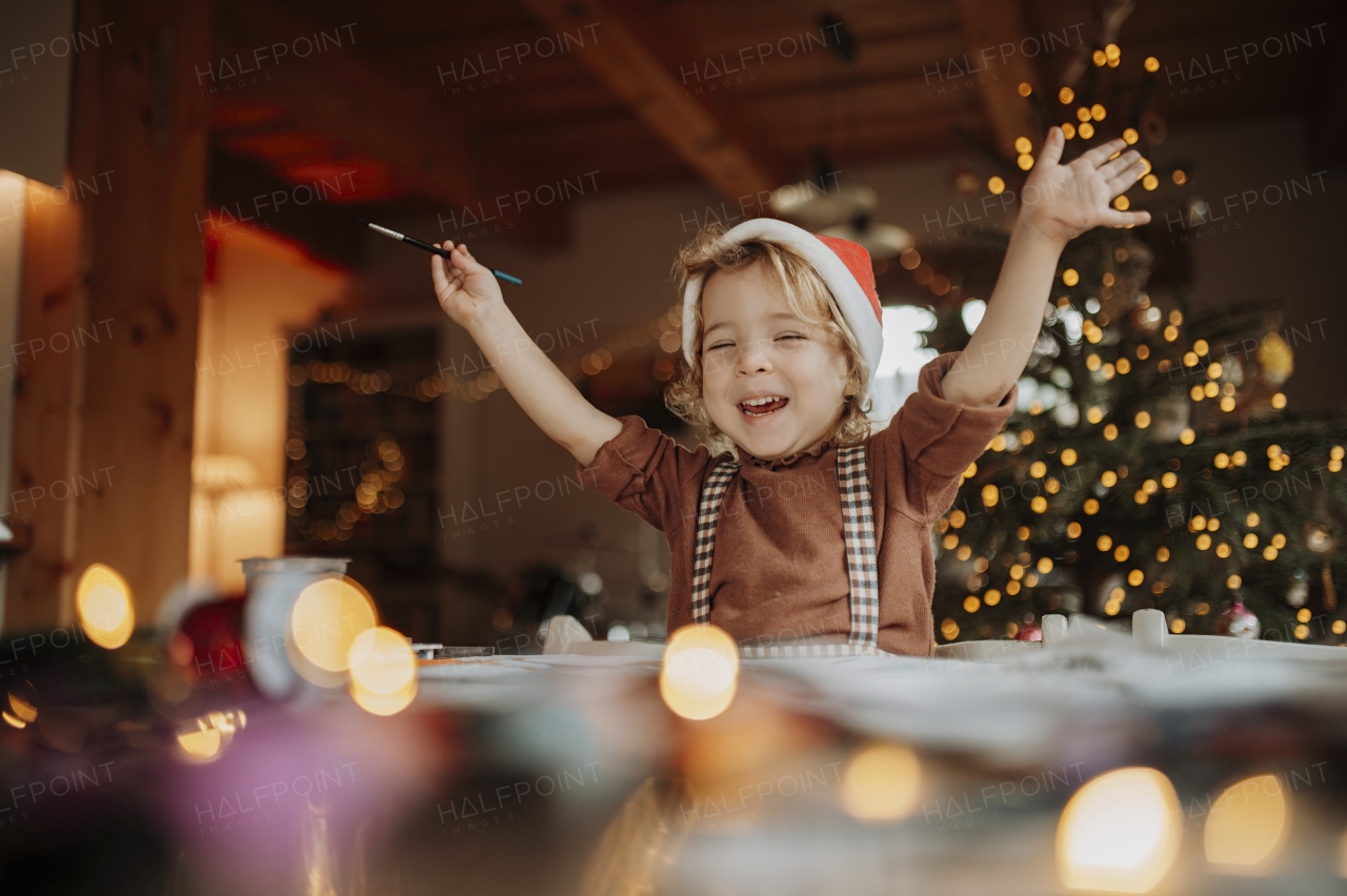 Portrait of small girl indoors at Christmas home, arms in the air. Cute girl painting picture for Santa Claus or making christmas decoration.