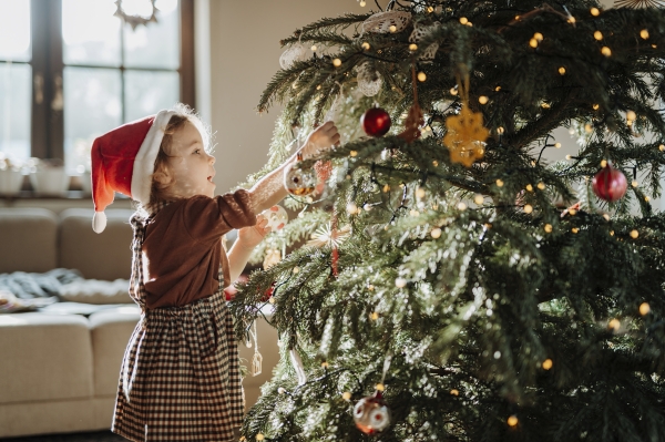 Portrait of cute girl in Santa's hat helping with decorating the Christmas tree with christmas ornaments.