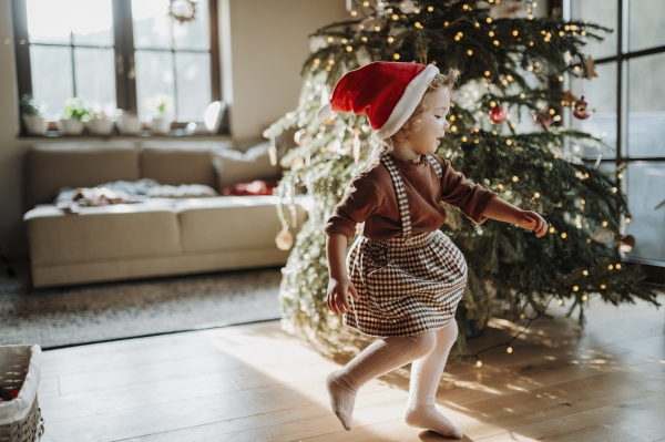 Portrait of cute girl in Santa's hat helping with decorating Christmas tree and dancing around the tree.