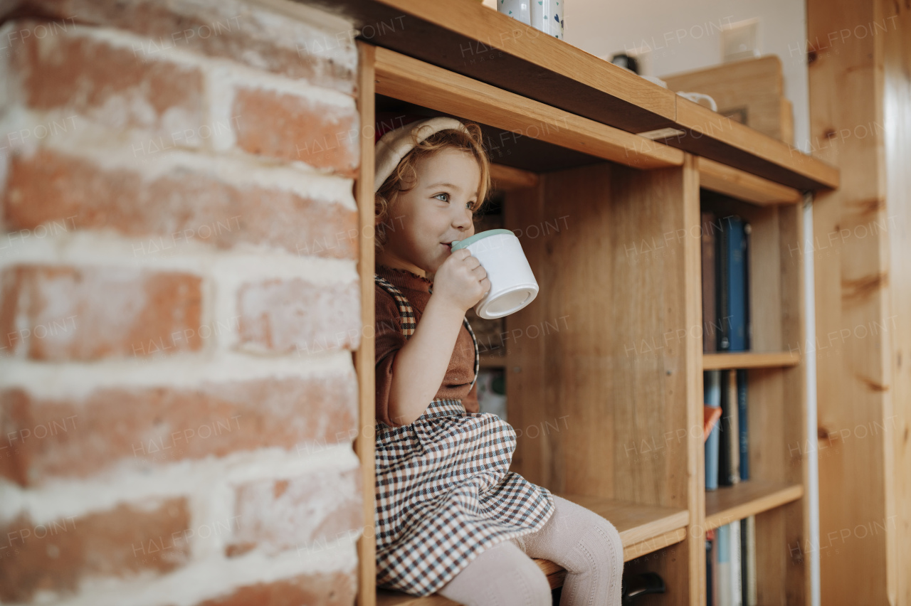 Cute little girl sitting in kitchen, drinking hot chocolate from mug, wearing a Santa hat. Festive atmosphere at home.