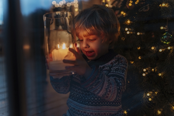 Cute little boy holding Christmas candle in his hands, Christmas tree behind him. Festive christmas atmosphere at home.