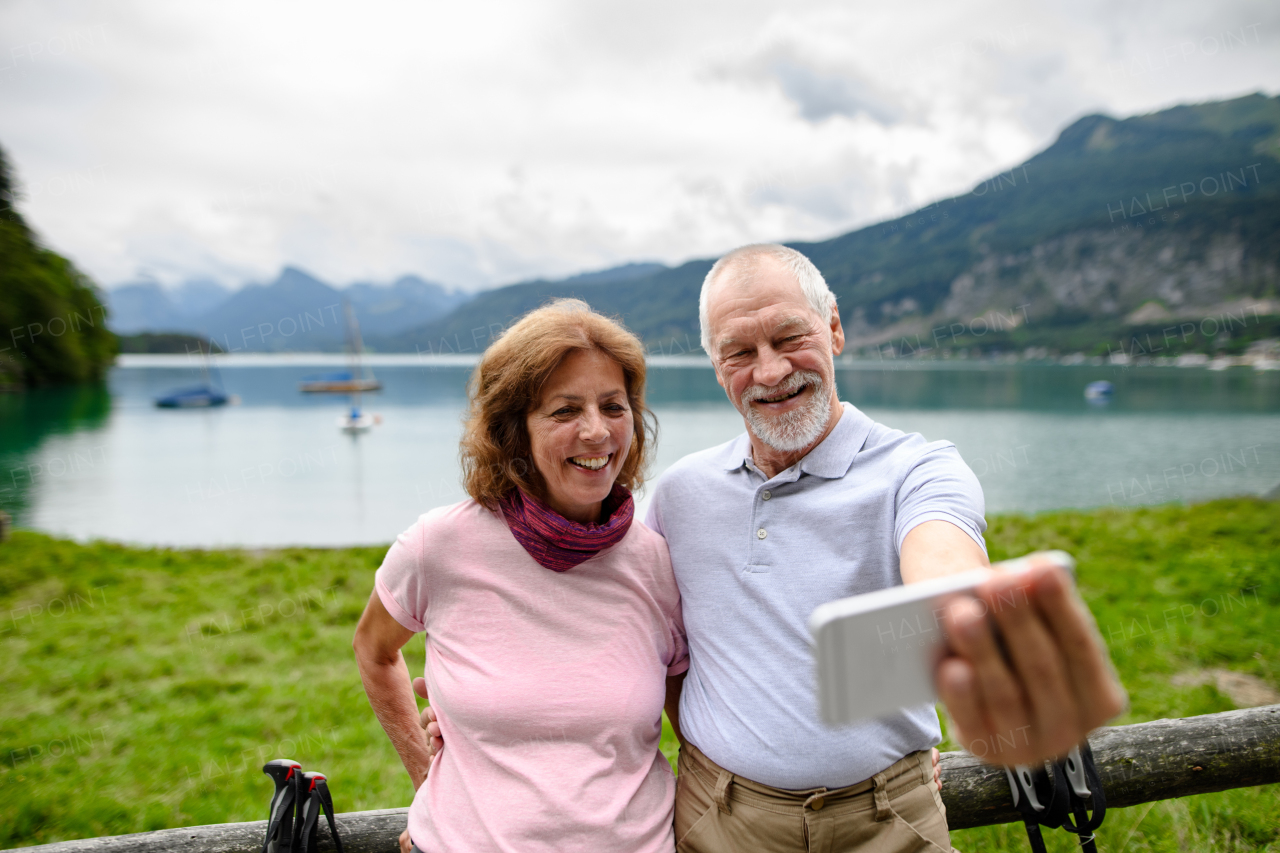 Portrait of elderly couple takiing selfie with smartphone while hiking together in mountains. Senior tourists embracing in front of lake.