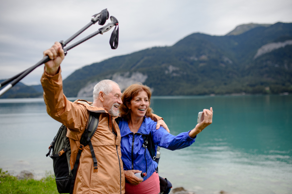 Portrait of beautiful active elderly couple taking selfie during hike in autumn mountains. Senior tourists embracing each other in front of lake.