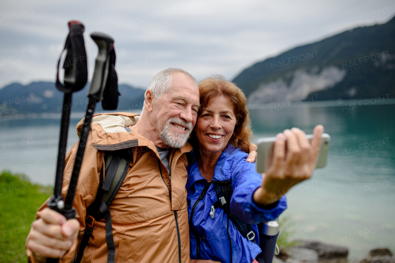 Portrait of beautiful active elderly couple taking selfie during hike in autumn mountains. Senior tourists embracing each other in front of lake.