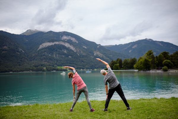 Senior couple doing outdoor yoga, tai chi, pilates by the lake in the autumn. Elderly husband and wife spending active vacation in the mountains. Pensioners stretching after outdoor workout.